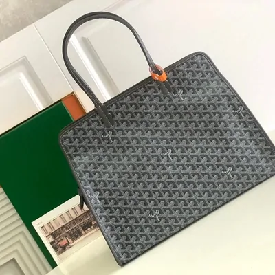 A grey and black chevron-pattern tote with a rolled leather handle, standing in front of its green box